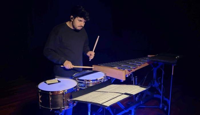 A musician dressed in black plays a marimba and two snare drums using drumsticks. 乐谱摊开在他面前的架子上.